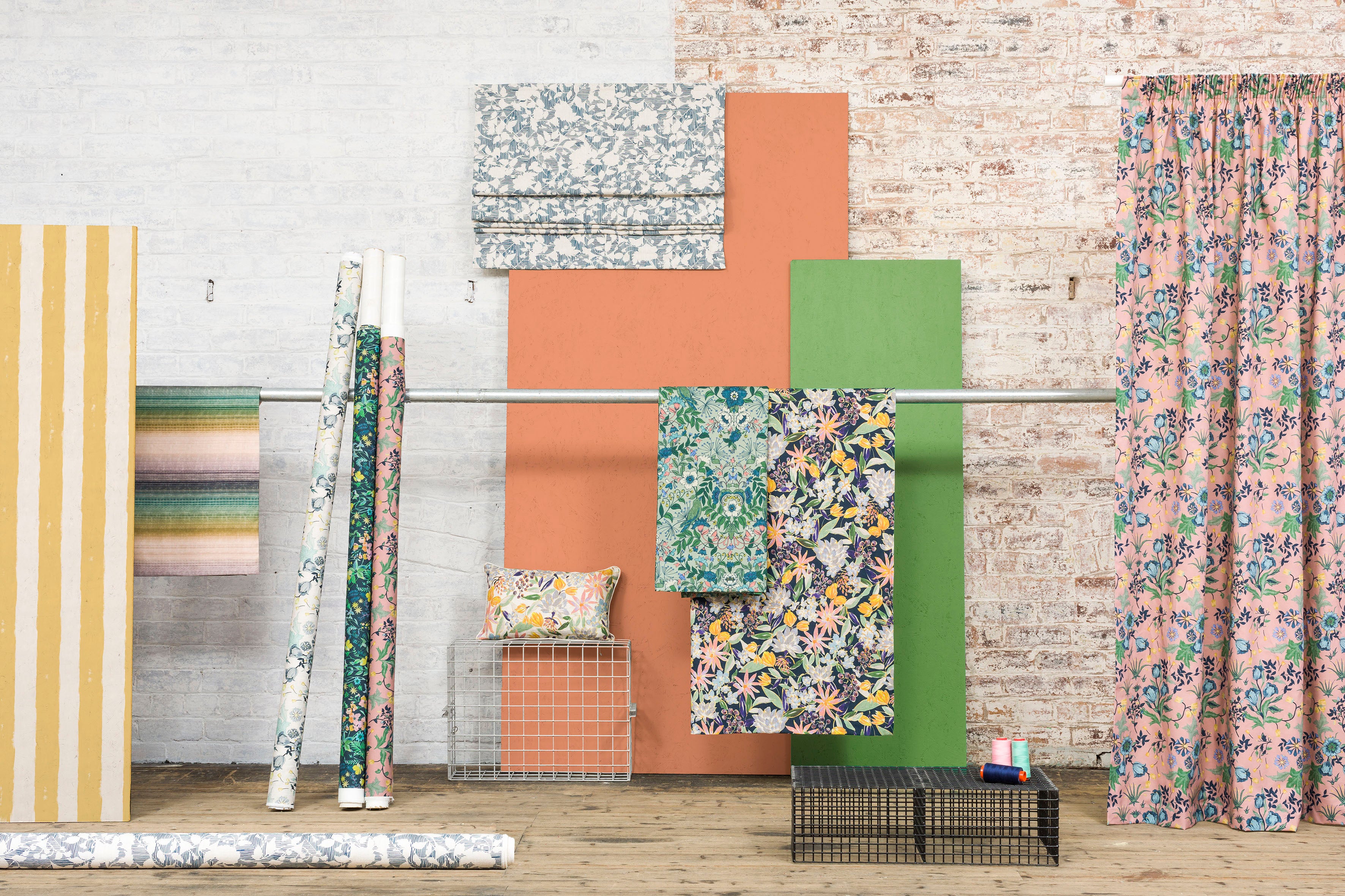 printed fabric shown in rolls of fabrics and displayed by hanging on a pole in a wide shot showing a range of designs agains a white brick wall